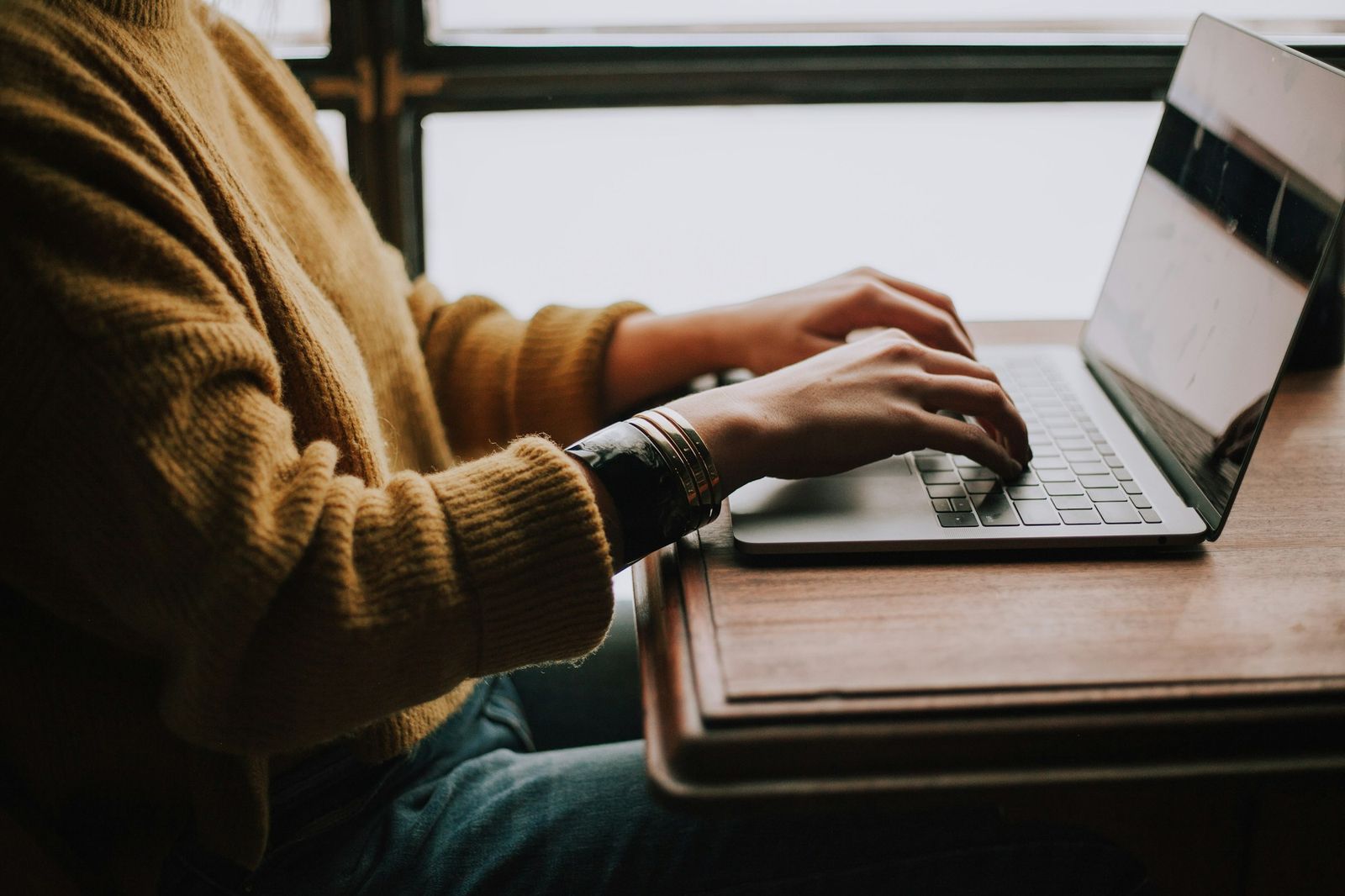 A person typing on a laptop at a wooden desk by a window