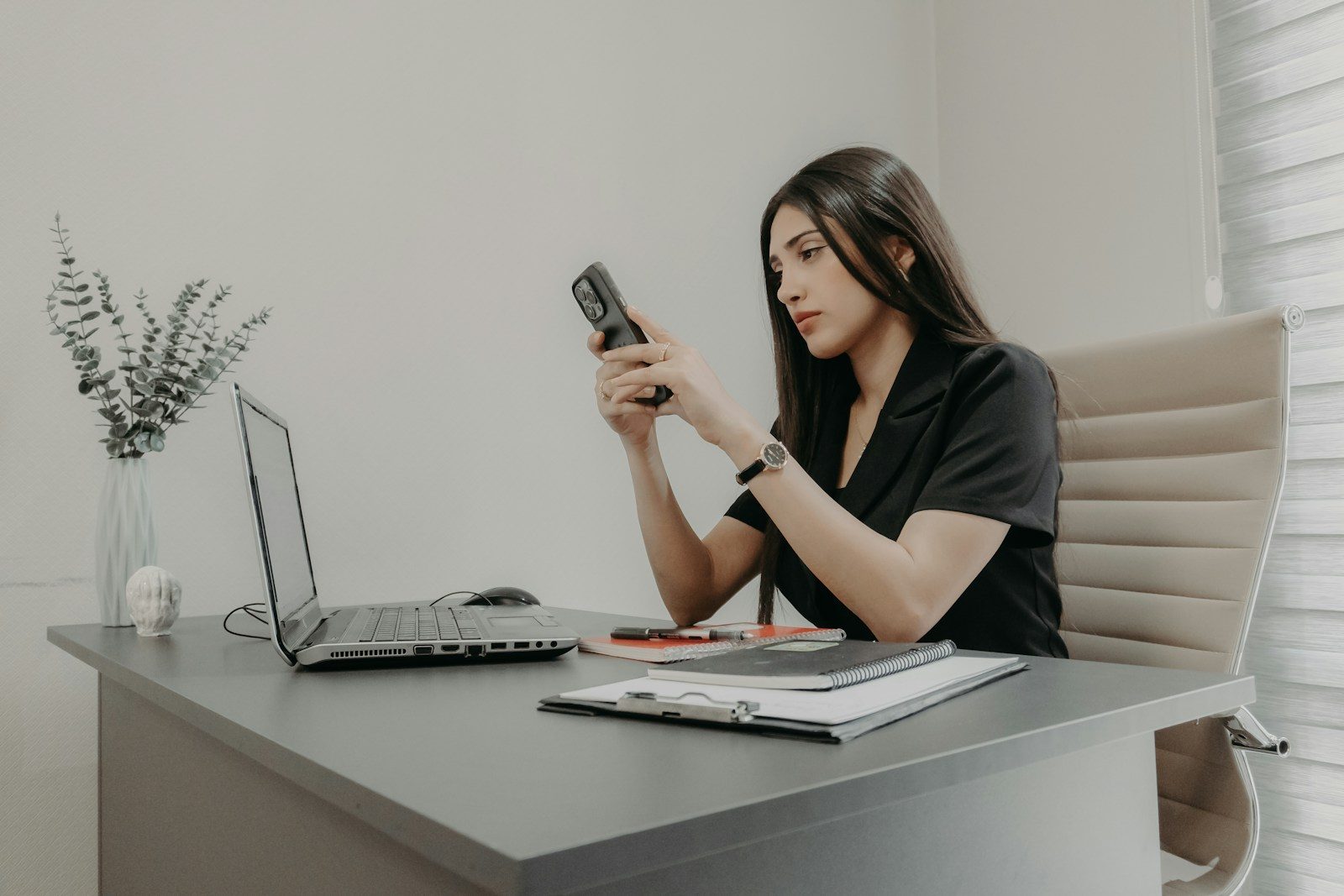A woman checking her phone at her desk, laptop open beside her
